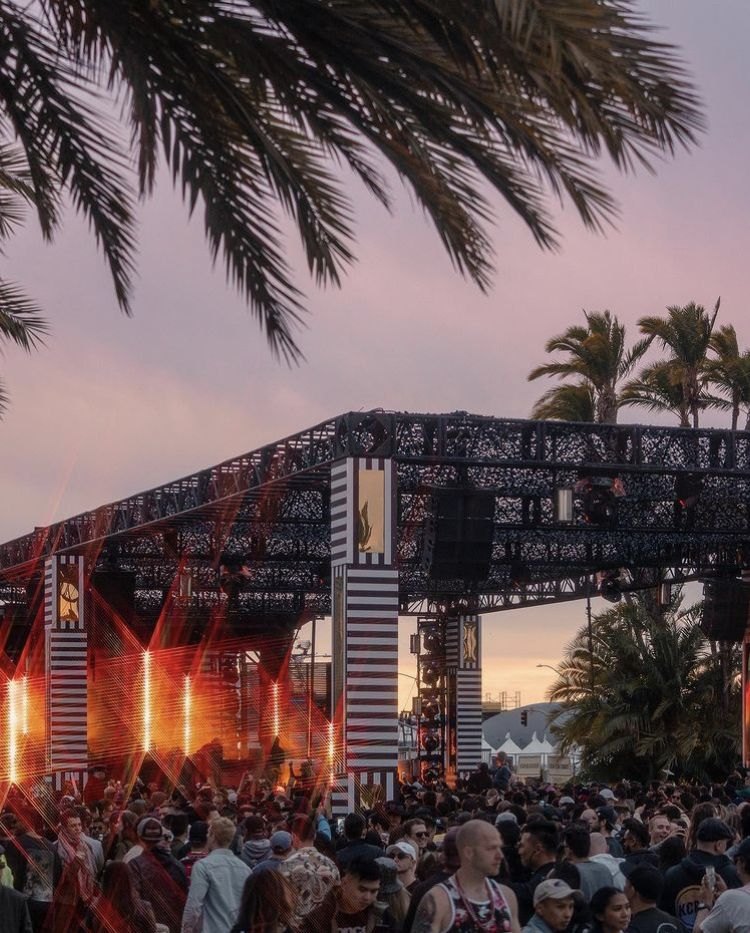 A festival main stage at sunset, lasers over a dense crowd, palm trees against a pink sky